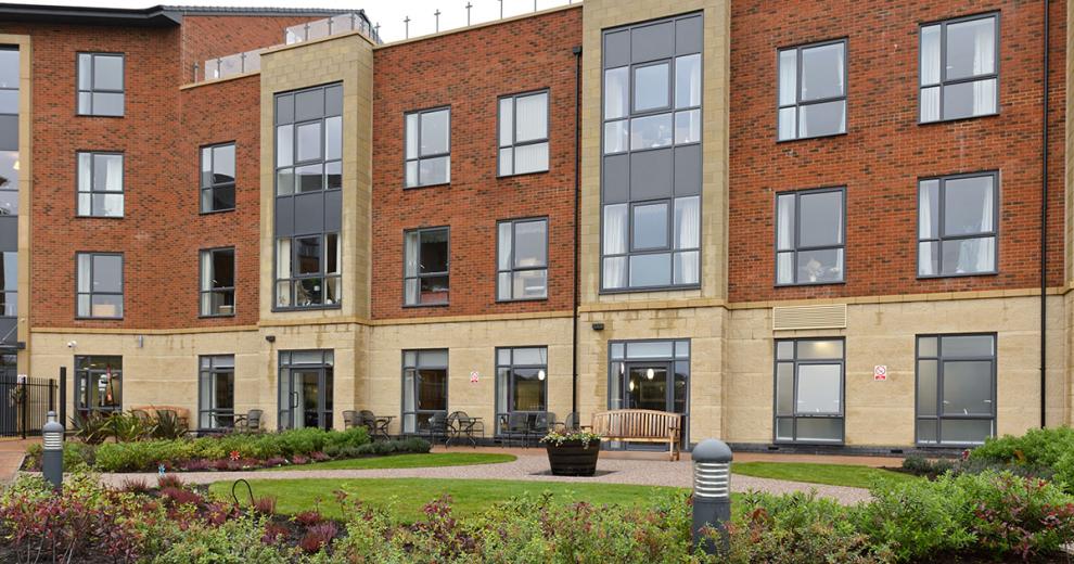 The four storey retirement community. The base of the property is built all in yellow brick, the further three storeys are built using a reddish brown brick with the protruding properties also built with the yellow bricks producing vertical stripes up the property. A outdoor rooftop balcony is seen on the right hand side of the property.