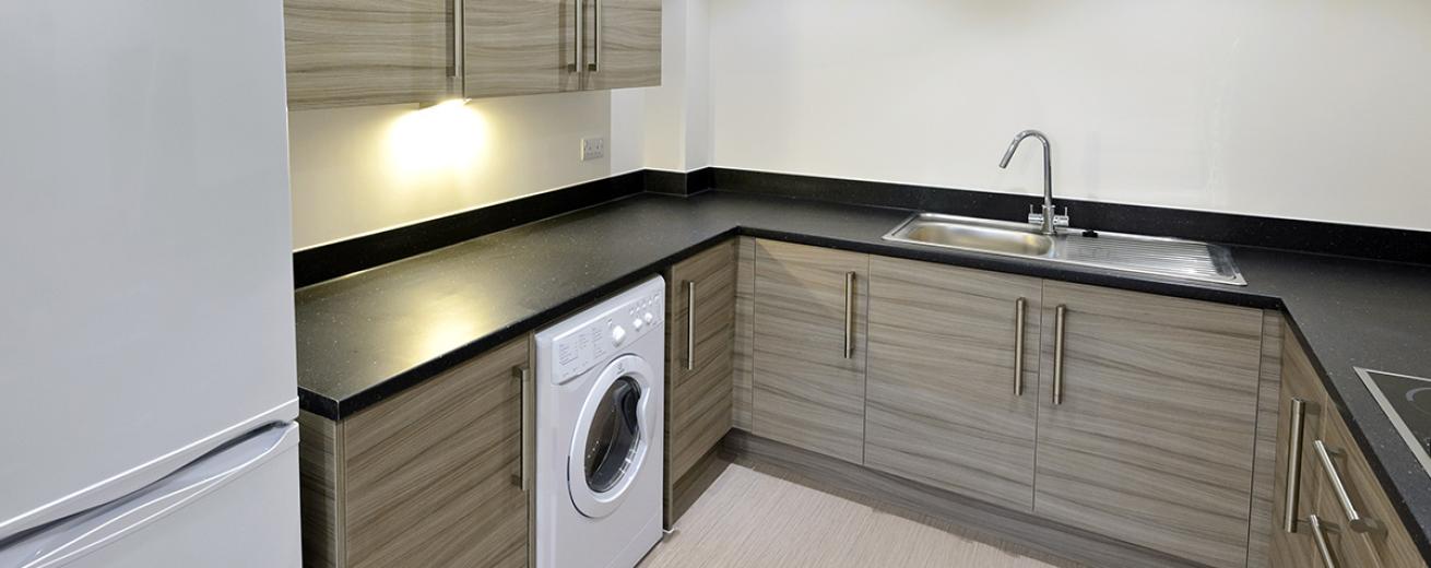 The kitchen has an earthy decoration using a midtone brown for the cabinetry, a stark black worktop with chrome sink. The washing machine and fridge freezer are a brilliant white.