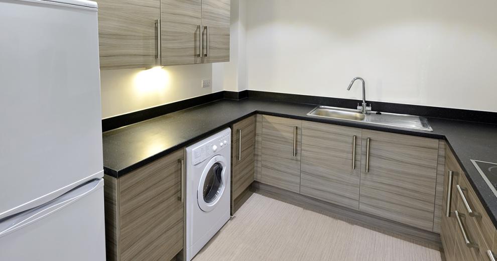 The kitchen has an earthy decoration using a midtone brown for the cabinetry, a stark black worktop with chrome sink. The washing machine and fridge freezer are a brilliant white.
