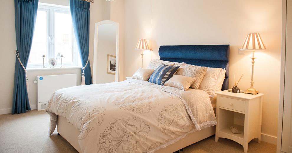 A tranquil minimal bedroom space, the navy double bed sits centrally on the back wall popping against the white décor. The navy curtains also add a calming colour to the room. The white bedside tables either side of the bed are adorned with tall thin lamps a full length white wooden framed mirror tucks neatly into the corner of the room.