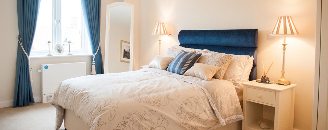 A tranquil minimal bedroom space, the navy double bed sits centrally on the back wall popping against the white décor. The navy curtains also add a calming colour to the room. The white bedside tables either side of the bed are adorned with tall thin lamps a full length white wooden framed mirror tucks neatly into the corner of the room.