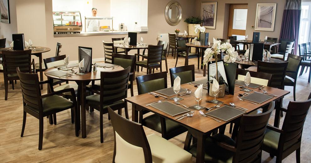 A sophisticated dining room of circular four person and rectangular six person wooden dining tables with black wooden chairs with a mixture of white and black leather upholstery, the tables are all dress with leather place mats cutlery, wine glasses with napkins and leather bound menus. All of the tables are placed in front of the industrial style dining hatch.