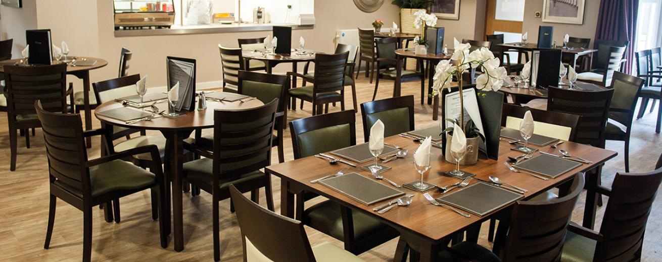 A sophisticated dining room of circular four person and rectangular six person wooden dining tables with black wooden chairs with a mixture of white and black leather upholstery, the tables are all dress with leather place mats cutlery, wine glasses with napkins and leather bound menus. All of the tables are placed in front of the industrial style dining hatch.