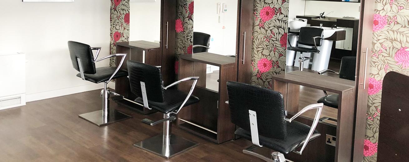 A minimalistic hair salon, three black leather and chrome styling chairs are placed in front of a small shelving unit with a full length mirror mounted into the unit. A fuchsia flower and buds amongst the green leaves wallpaper design behind the mirrored units.