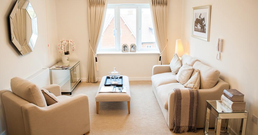 Beautiful bright and airy living area with cream carpets, light beige three seater sofa and matching arm chair and footstool, dressed with a black tray, a decanter and glasses. Mirrored furniture is used as a decorative piece in the corner of the room and mirrored small coffee table is placed next to the sofa with decorative books.