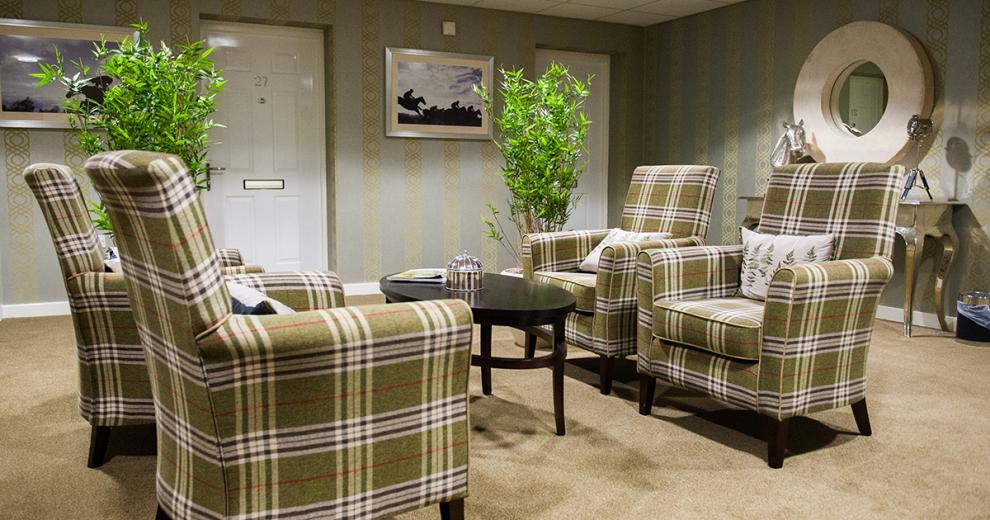 Four green tartan patterned tall backed arm chairs, two placed either side of the of a black oval coffee table. The room is decorated in a khaki green wallpaper with a lime green vertical pattern design. Two large potted plants with small trees sit at the door entranceway and adjacent to two of the arm chairs.