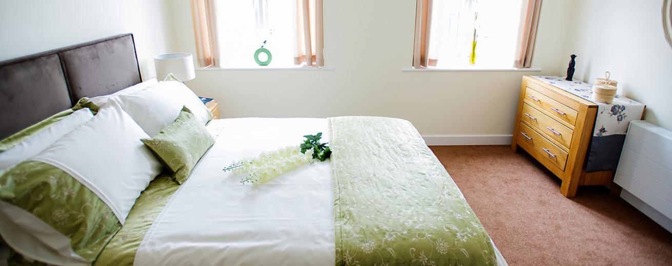 A clean and fresh double bedroom, home to a large double bed placed centrally in the room, decorated in white bedding with a green and white floral embroidery. The room benefits from two large windows which fill the majority of the wall flooding in the natural light. A wooden chest of draws its tucked neatly into the corner of the room between the window and the radiator.