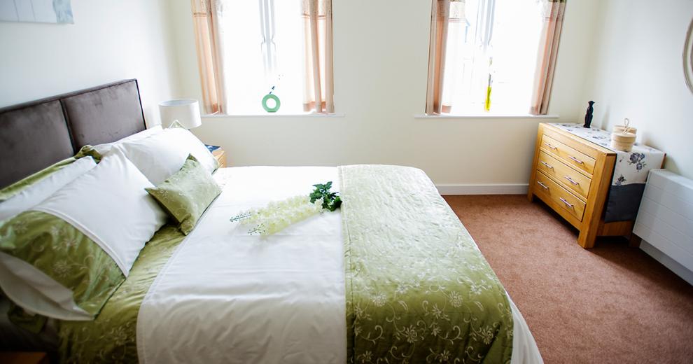 A clean and fresh double bedroom, home to a large double bed placed centrally in the room, decorated in white bedding with a green and white floral embroidery. The room benefits from two large windows which fill the majority of the wall flooding in the natural light. A wooden chest of draws its tucked neatly into the corner of the room between the window and the radiator.