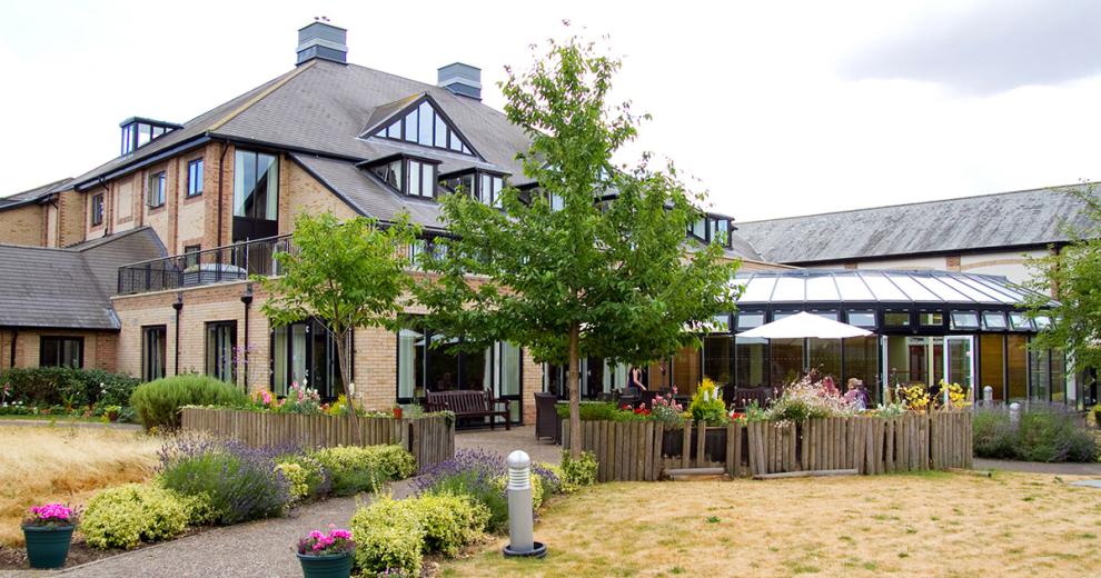 An idyllic brick built property with mass amounts of over sized windows and added skylights, set amongst a brick paved patio area and parasol seating enclosed in by wooden logged planters and colourful plants.
