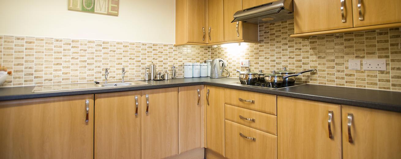 A fully functional kitchen with lightly stained wooden cabinetry on the lower and upper units, a black worksurface, an electric hob and chrome accessories.