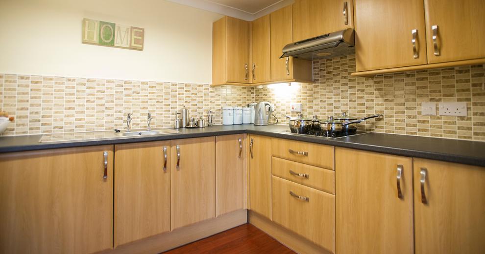 A fully functional kitchen with lightly stained wooden cabinetry on the lower and upper units, a black work surface, an electric hob and chrome accessories.