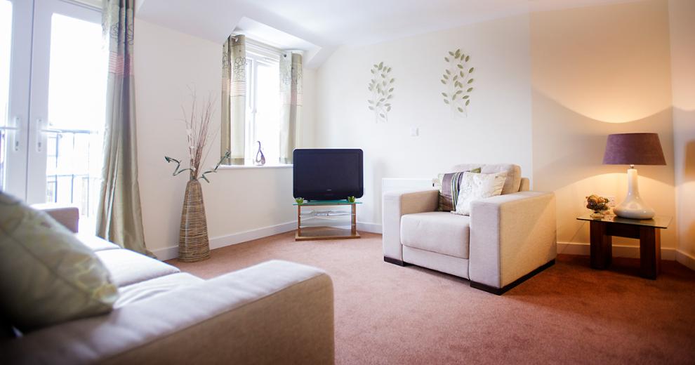Crisp and fresh living room with a cream boxy three seater cotton furniture and matching armchair. A petite coffee table sits in the corner of the room next to the arm chair with a large ceramic lamp and purple lamp shade, the television and glass television stand sit the other side of the arm chair. In front of a large window draped in short green curtains. Two patio doors open out onto a balcony area draped in the same curtains but in full floor length as the window.