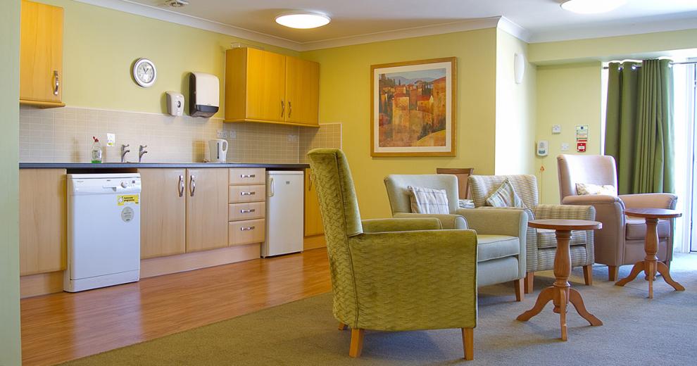 A pastel green decorated seating area, multiple cushioned armchairs facing the far end of the seating area. Behind is a neat kitchenette area with dishwasher, undercounter fridge, multiple wooden cabinets, a sink and tea making facilities.
