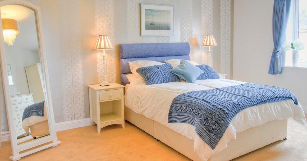 A serene double bedroom, the blue double bed sits centrally on the back wall popping against the white décor. The matching blue curtains also add a calming colour to the room. The white bedside tables either side of the bed are adorned with tall thin lamps a full length white wooden framed mirror tucks neatly into the corner of the room.