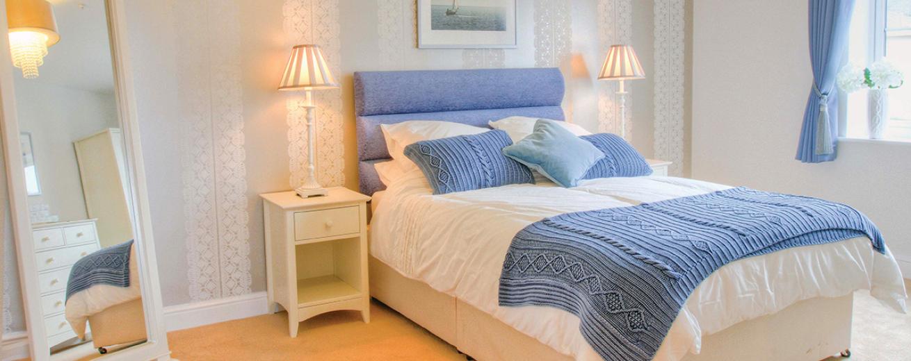 A serene double bedroom, the blue double bed sits centrally on the back wall popping against the white décor. The matching blue curtains also add a calming colour to the room. The white bedside tables either side of the bed are adorned with tall thin lamps a full length white wooden framed mirror tucks neatly into the corner of the room.