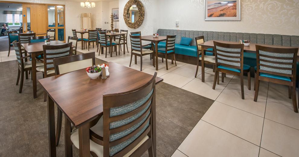 The spacious dining room hosts booth and dining chair seating options for the residents. The dark wooden stained dining tables and matching chairs the chairs are upholstered with teal and cream seat cushions. The booth seating is decorated in teal and silver fabrics. The dining room links through into the seating area.