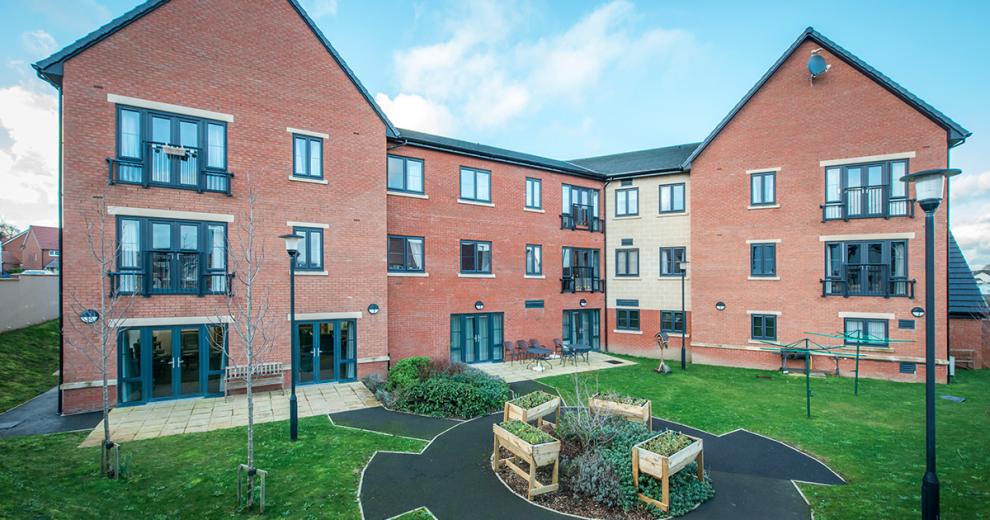 A modern red bricked new build retirement property. The building is built as a three storey corner plot with a small patio area along the left hand side of the property hosts an array of seating options from a wooden bench or metal chairs and circular tables. The patio leads into a circular tarmacked area with wooden planters and shrubbery, the rest of the garden is covered in grass.