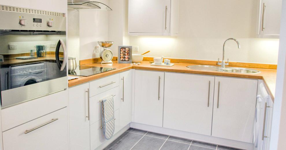 A beautifully airy white kitchen, featuring white cabinetry with under lighting adding more brightness against the white painted walls. The mounted wall oven, backsplash and extractor fan and circular sink are all in chrome which reflects the light perfectly. The room is warmed by a wooden kitchen counter surrounding the room, broken up by the electric hob and baking accessories used as decorations.