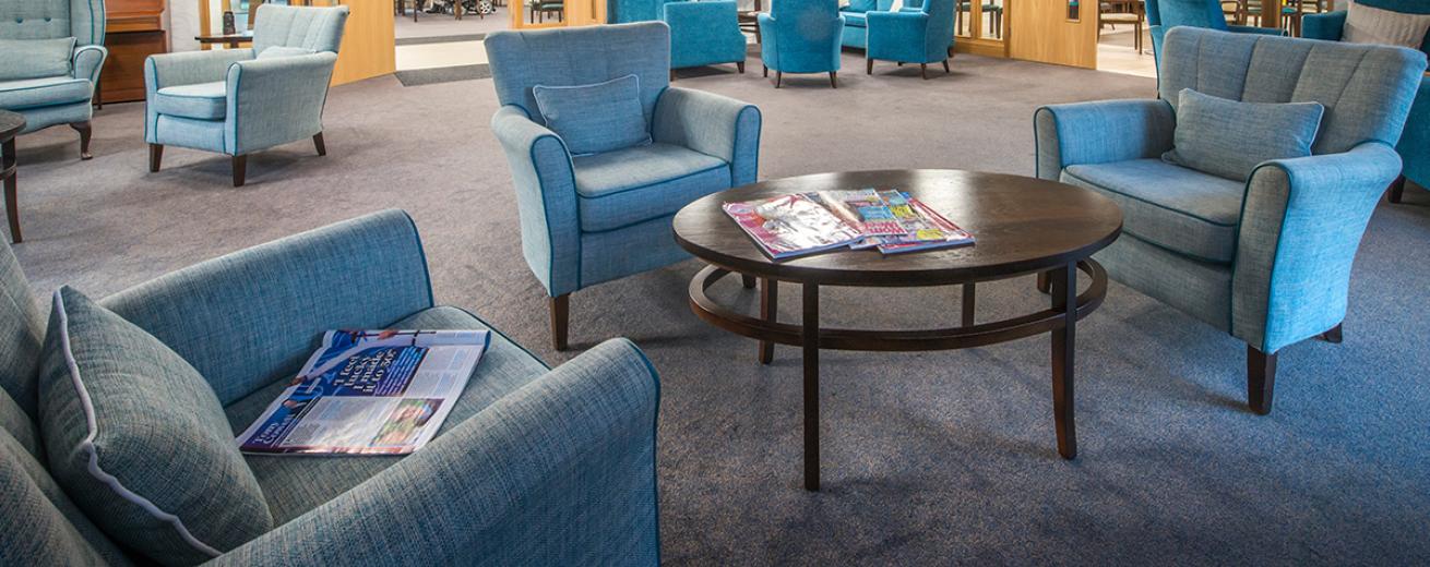 A large and brightly blue dressed communal seating area. The array of different shades of blue armchairs placed in small clusters circling an oval mahogany coffee table.