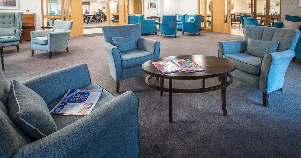 A large and brightly blue dressed communal seating area. The array of different shades of blue armchairs placed in small clusters circling an oval mahogany coffee table.