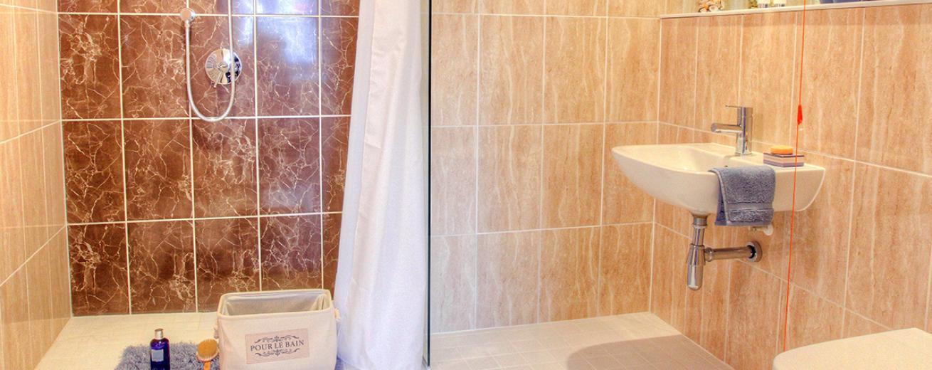 The shower room is decorated in oversized wall tiles the majority are a soft brown with white veining, the panel behind the shower is decorated in a bronzed, white vein tile. The room is minimal with a wall mounted white toilet and sink basin.
