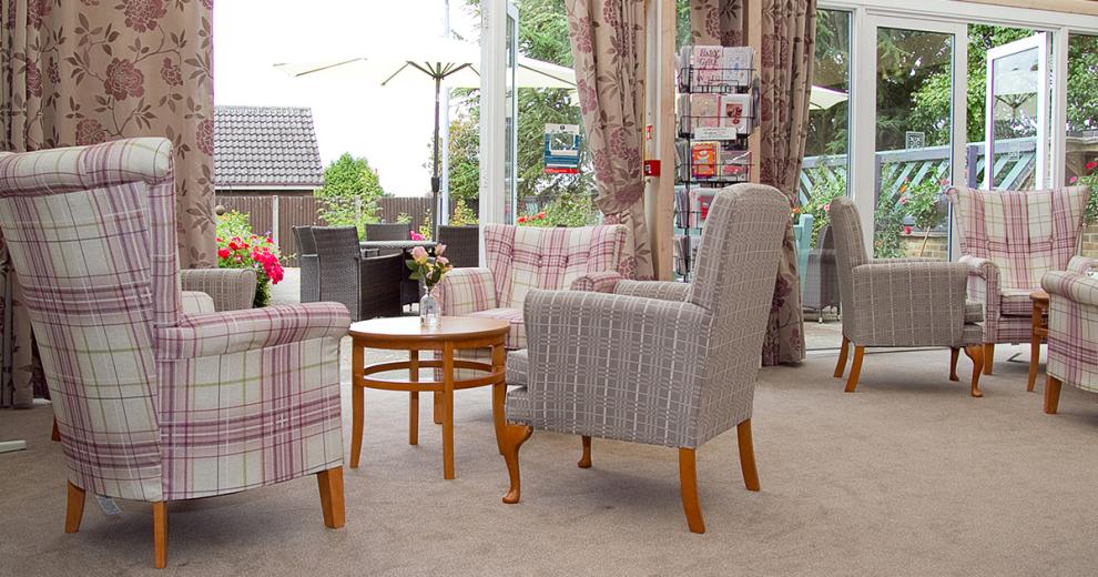 The seating area is surrounded by patio doors leading out into the garden and out door seating area. The plush grey and purple and white checked armchairs are placed around a small wooden circle coffee table facing out towards the outdoors.