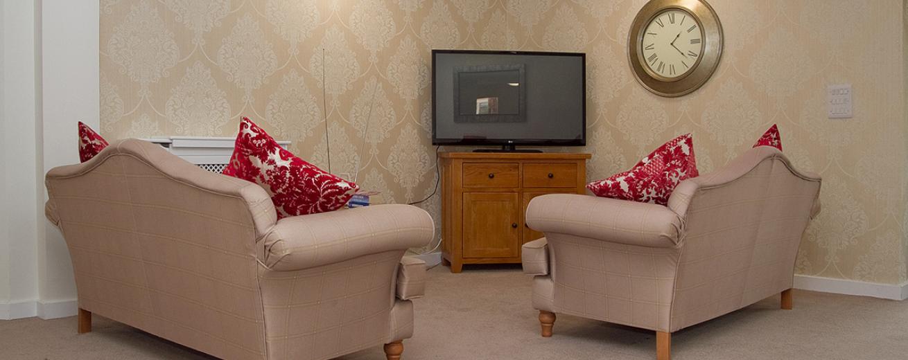 The television nook is an intimate seating area with two ornate two person sofas facing into the television placed onto of a midsized wooden television unit. The cream fleur de lis wall paper adds warms and a cosy feel to the corner.