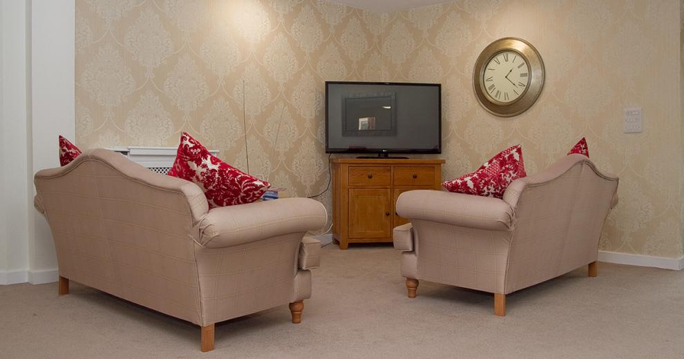 The television nook is an intimate seating area with two ornate two person sofas facing into the television placed onto of a midsized wooden television unit. The cream fleur de lis wall paper adds warms and a cosy feel to the corner.