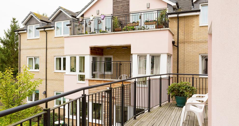 The thin but long balcony spans the with of the private residence. With black iron railings and a wooden decked flooring. With two chairs that overlook the garden area of the property.