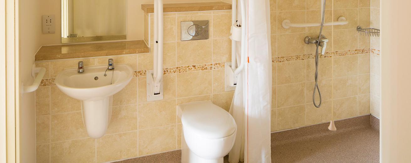 The cream and white veined tiled bathroom with walk in shower and wall mounted white toilet and skink with a mirror placed centrally above the sink butted up to the shelf.