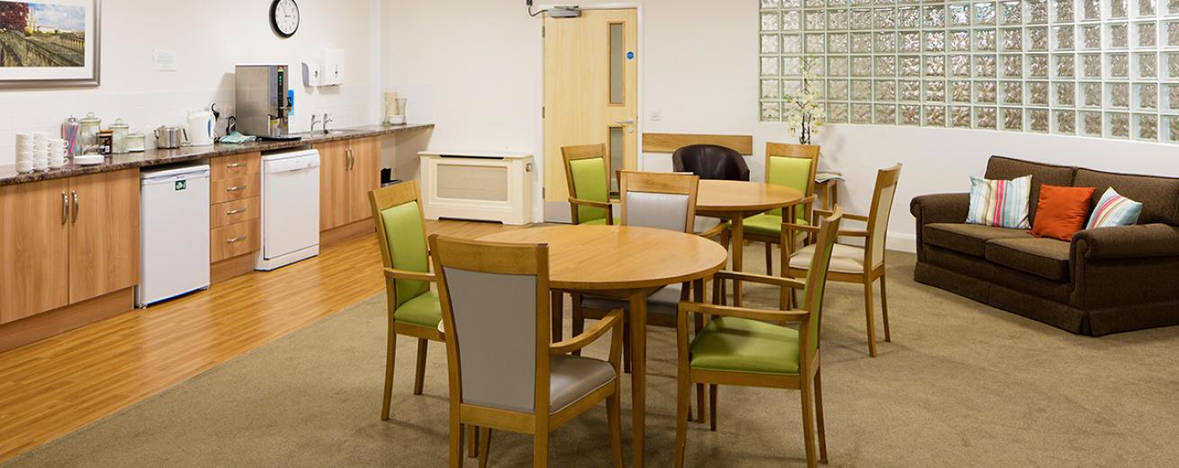 A large kitchenette spans the width of the back wall of the seating area. The soft wooden cabinets and black worktops complement the white room well. The kitchen features a dishwasher and undercounter fridge as well as numerous tea and coffee making facilities. Two four seater dining tables with lime green and lilac chairs are placed in front of the kitchen. A small black sofa tucks underneath the mottled glass panelled across the wall.