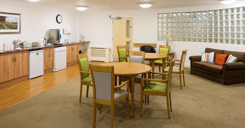 A large kitchenette spans the width of the back wall of the seating area. The soft wooden cabinets and black worktops complement the white room well. The kitchen features a dishwasher and undercounter fridge as well as numerous tea and coffee making facilities. Two four seater dining tables with lime green and lilac chairs are placed in front of the kitchen. A small black sofa tucks underneath the mottled glass panelled across the wall.