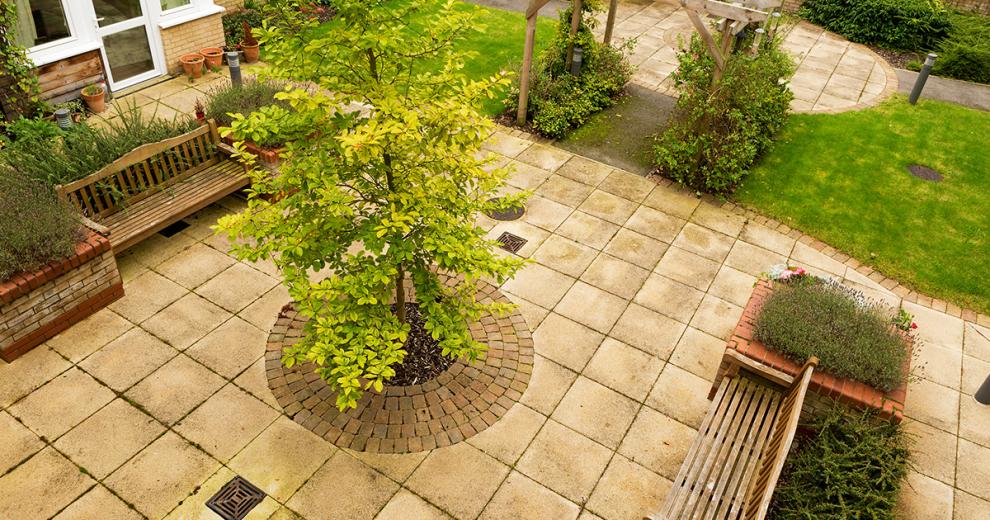 The majority of the courtyard is paved with small grassed patches framing the edging. The use of trees, shrubbery and potted plants make the space feel very fresh. Two large benches sit around the tree and amongst the shrubs.