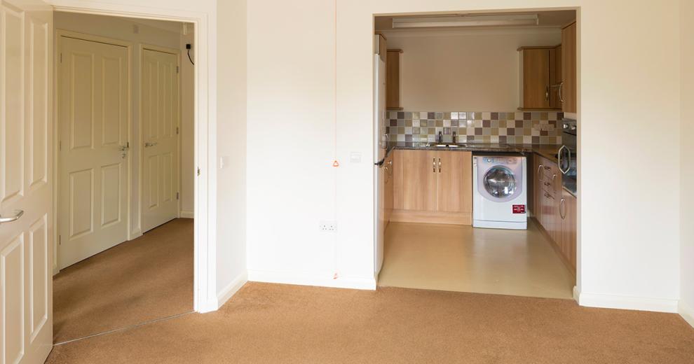 Crisp and fresh living area, leading through into a fully functional kitchen with lightly stained wooden cabinetry on the lower and upper units, a black work surface, an electric hob, washing machine and chrome accessories.