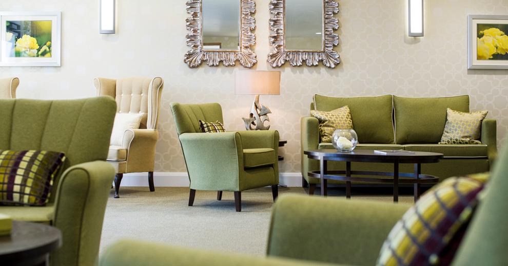 A sophisticated communal seating area. Displaying a soft cream patterned design feature wall. The dark wooden oval coffee table and circular side table complement the olive green sofas and armchairs. Bold ornate mirrors adorn the walls and artistic sculpture lamps add soft lighting.