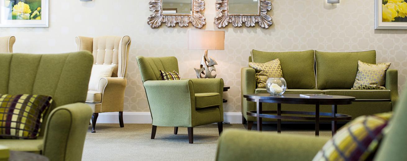A sophisticated communal seating area. Displaying a soft cream patterned design feature wall. The dark wooden oval coffee table and circular side table complement the olive green sofas and armchairs. Bold ornate mirrors adorn the walls and artistic sculpture lamps add soft lighting.
