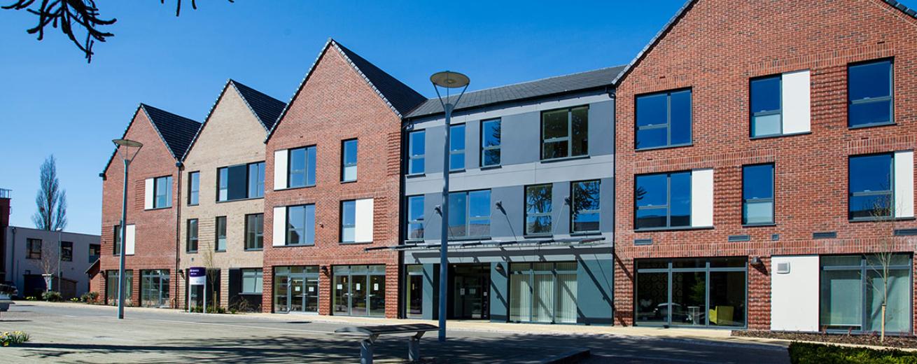 New build red brick built three storey complex, with exaggerated triangular roofing. The entrance way is a grey slopped roof design nestled between two of the buildings.