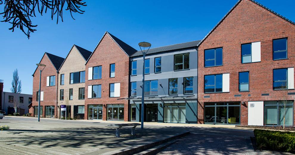 New build red brick built three storey complex, with exaggerated triangular roofing. The entrance way is a grey slopped roof design nestled between two of the buildings.