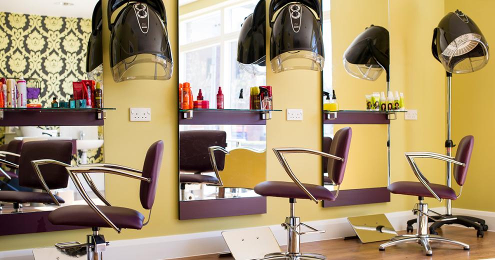A warmly designed hair salon area. Three floor length elongated mirrors are the main feature of the room. Three purple styling chairs are located in front of the mirror, with modern hair care products displayed on the shelves in front of the mirrors.