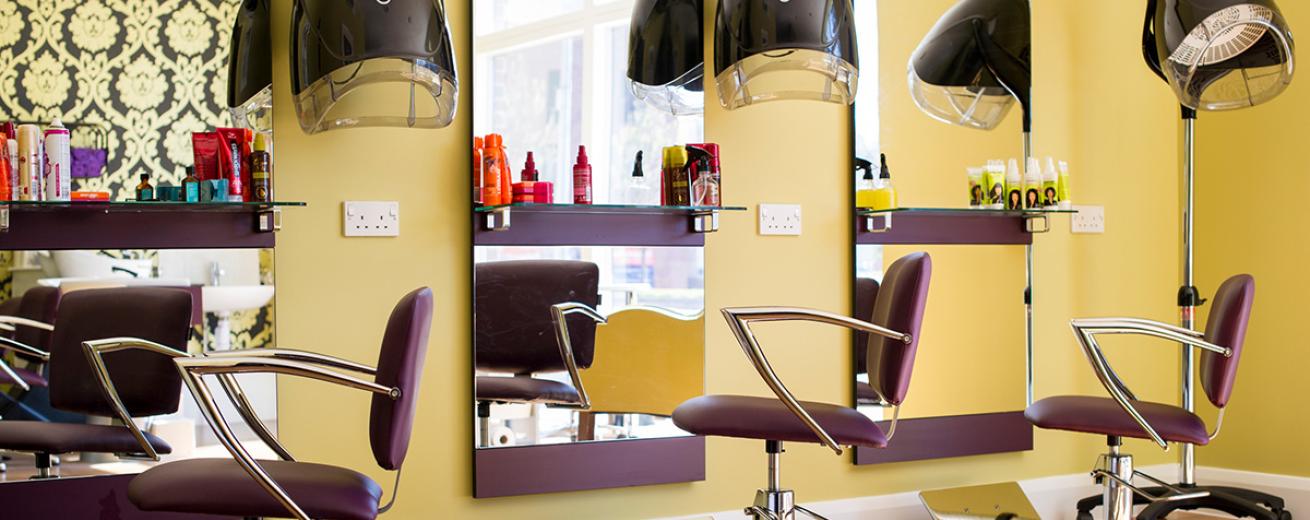 A warmly designed hair salon area. Three floor length elongated mirrors are the main feature of the room. Three purple styling chairs are located in front of the mirror, with modern hair care products displayed on the shelves in front of the mirrors.
