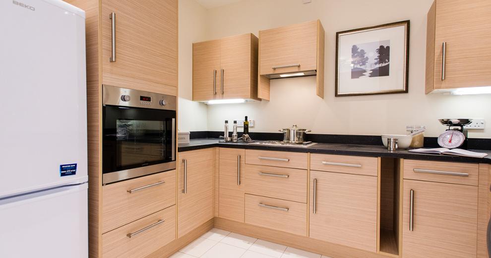 A compact white kitchen, with beach wood cabinetry with white painted walls decoration. The wall mounted oven and sink are all in chrome reflects light from the under-cabinetry lighting. The room is finished with a black work surface and brilliant white fridge freezer.