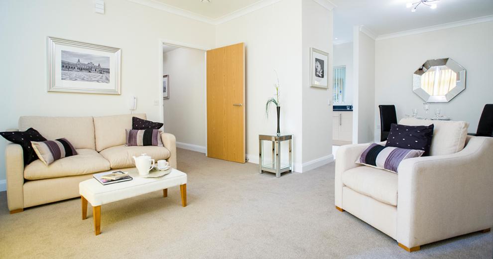 The generous living room is decorated neutrally with a two-seater cream fabric sofa. The matching armchair sits opposite the sofa separated by a footstool being used as a coffee table with tea making facilities ready for use. A hexagonal mirror sits above the two person black leather dining table in the nook of the room leading into the kitchen area.