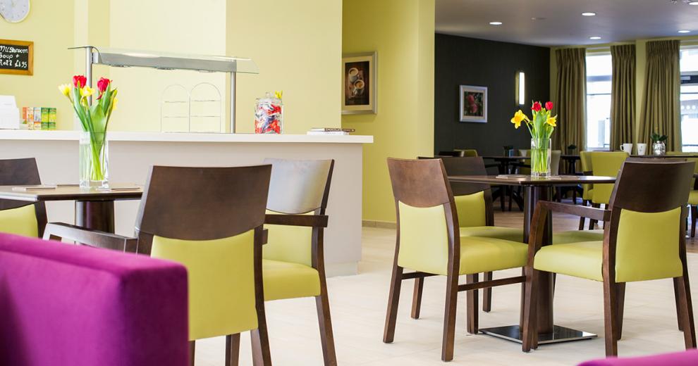 An enormous restaurant area. Pale yellow and sark green painted walls separate the areas. A vast selection of mahogany circular dining tables and chairs with lime green upholstery surround both areas. Amongst the yellow area is the industrial style serving hatch with heat lamps.