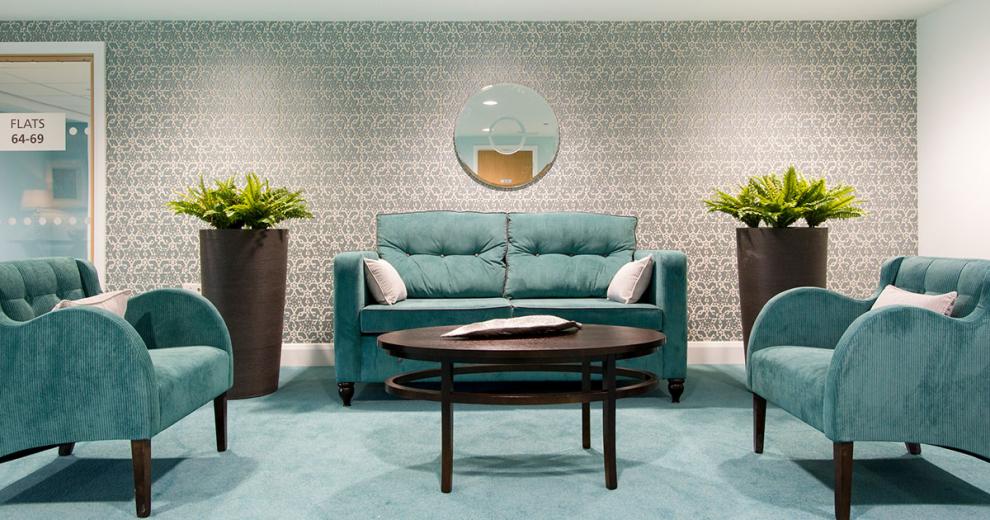 Bold teal seating area with plump crushed velvet two seater sofa and two arm chairs arranged around a large oval coffee table with two smaller circular tables placed either side of the sofa. The teal carpet mates the seating décor and contrasts with the earth toned, fleur de lis wallpaper.