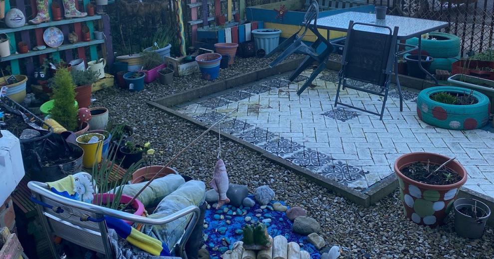An eclectic front garden mix, housing a slightly sunken paved outdoor seating area with blue painted mosaics. Surrounded by stone shingles and an array of multicoloured potted planters.