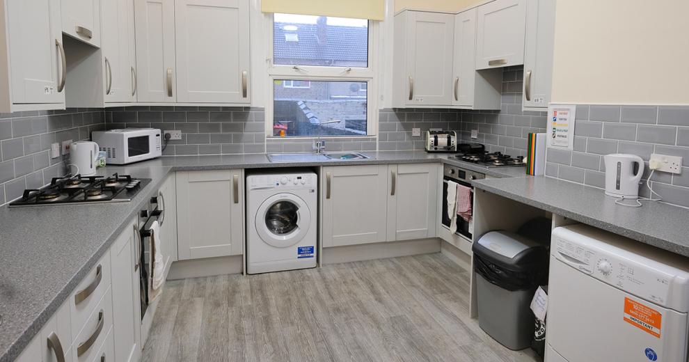 A modern grey toned kitchen. The light silvery grey painted cabinetry, grey mottled worksurface, a midtoned grey subway tile around all three walls. As well as housing two gas ringed hobs, two electric ovens, a white dishwasher and washing machine. A large window sits over the sink overlooking the garden.
