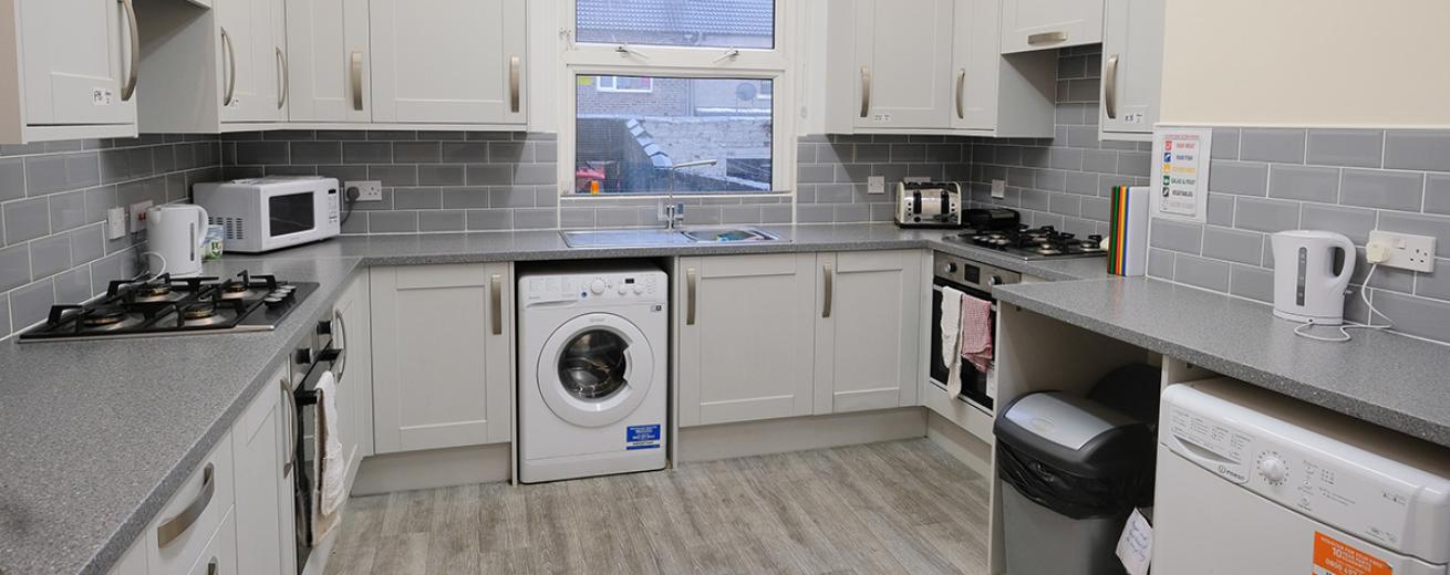 A modern grey toned kitchen. The light silvery grey painted cabinetry, grey mottled worksurface, a midtoned grey subway tile around all three walls. As well as housing two gas ringed hobs, two electric ovens, a white dishwasher and washing machine. A large window sits over the sink overlooking the garden.