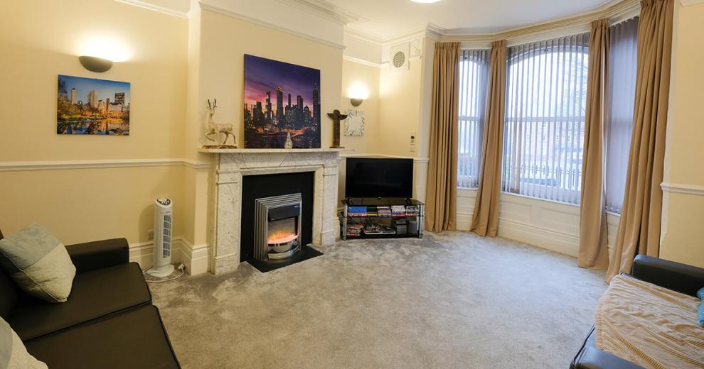 A cosy seating area featuring a large bay window. Two two-seater black leather sofas decorated with soft cream throwovers. Face towards the marble effect fireplace and electric fire and the television.