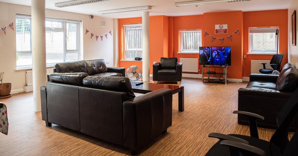 A good sized seating area with one bold orange painted feature wall with the television in front of it. Three black leather sofas, a black leather arm chair and two black wheeled computer chairs are circled around the television with a high gloss black coffee table centrally in the middle of them.