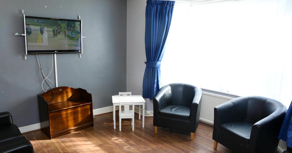 Tasteful grey decorated lounge with two black leather bucket armchairs under a wall sized window. The room is minimal with a wall mounted television and television unit underneath.